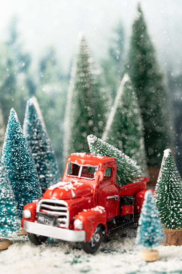 A photo of a traditional red toy truck carrying some toy christmas trees in the snow.
