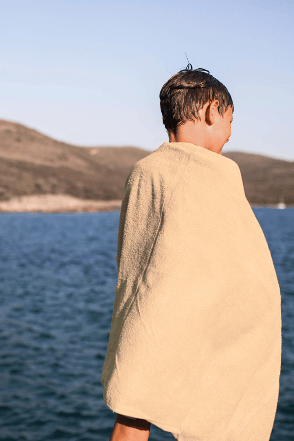 A young boy stands facing away from the camera with a brown towel wrapped around his shoulders looking out to sea. 