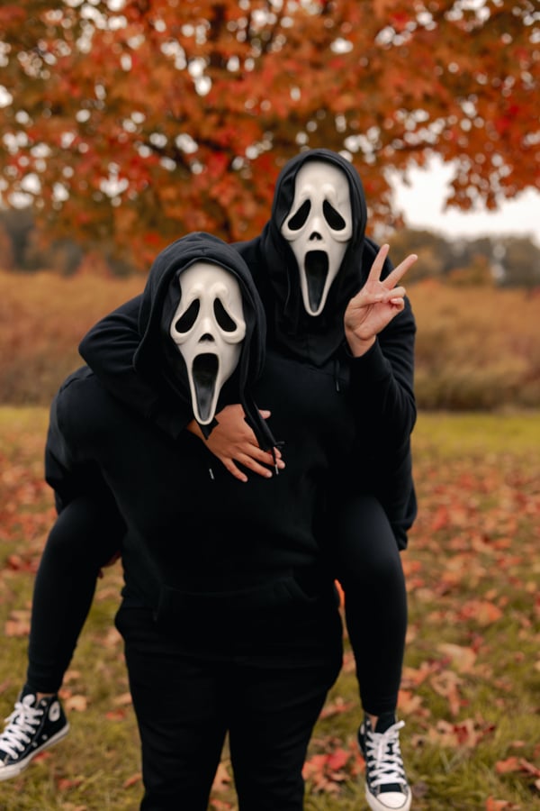 Two people wearing Scream masks playing around in an autumnal field.