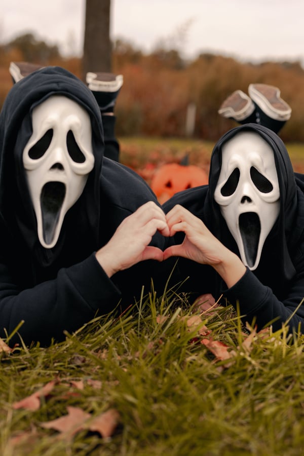 Two people lying next to eachother with Scream masks on making a heart-shape with their hands. 