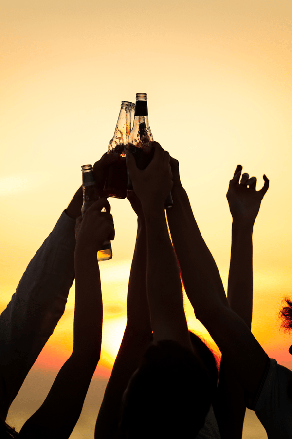 A group of people silhouetted against the sun holding up drinks to cheers. 