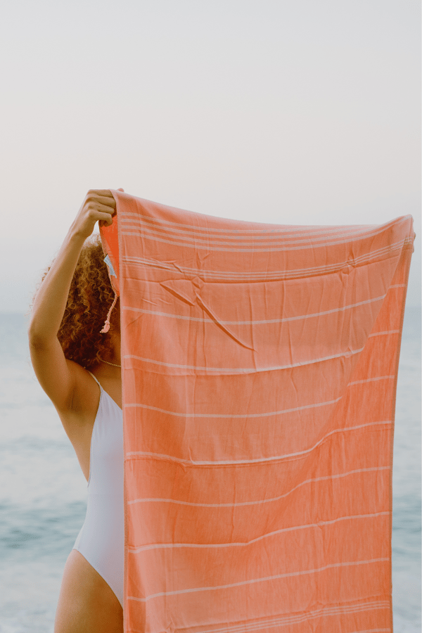 Someone holding up an orange beach towel on the beach. 