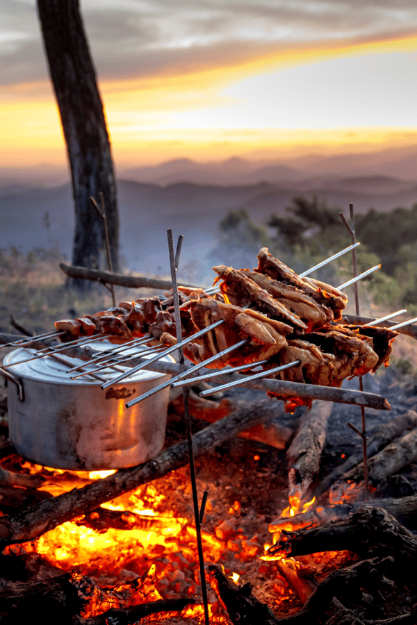 A BBQ cooking some meat skewers on an open fire at a campsite. 