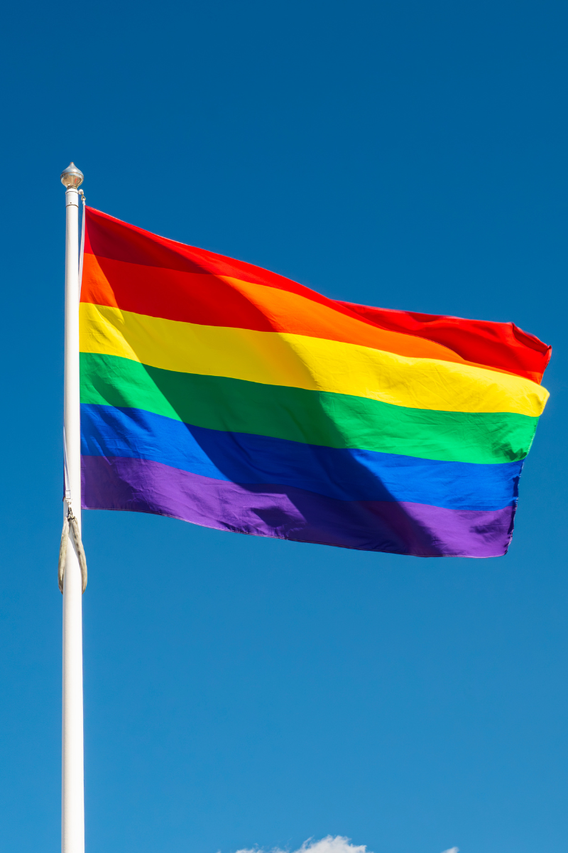 A Pride rainbow flag in front of a bright blue sky.