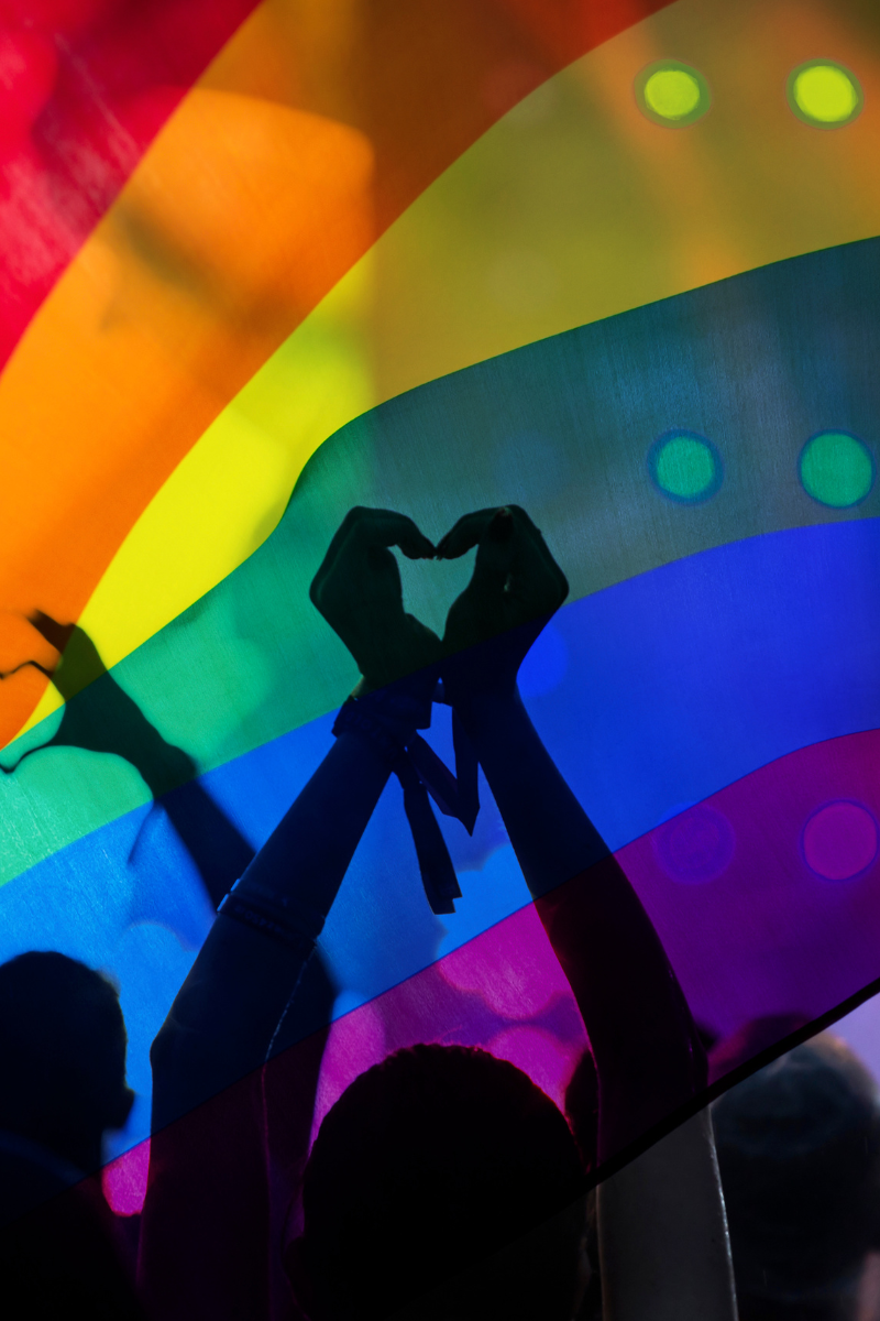 The silhouette of a person making the sign of a heart with their hands in front of a Pride rainbow flag.