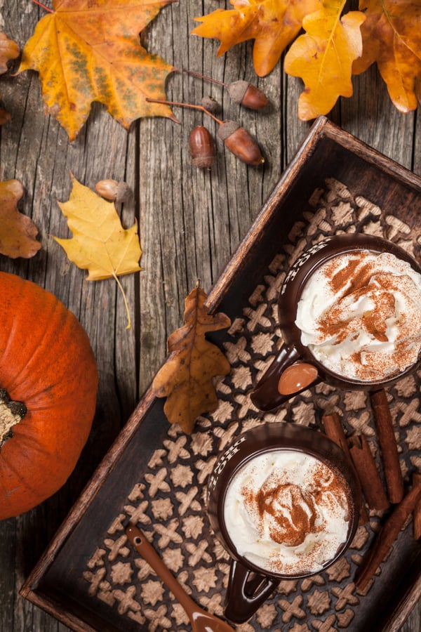 Two PSLs (Pumpkin Spice Lattes) on an autumnal tray, surrounded by some autumn leaves and pumpkins.