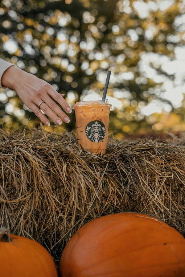 A Starbucks PSL (Pumpkin Spice Latte) sitting on a hay barrel behind some pumpkins.