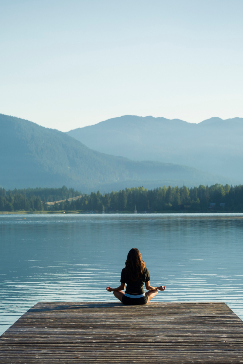 someone meditating on a wooden pier that's looking out over a quiet lake surrounded by trees for wellness and tranquility.