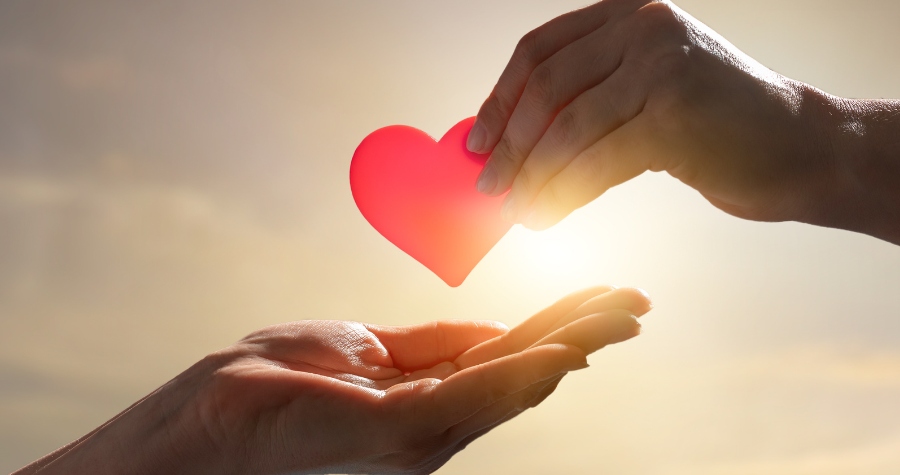 Two hands holding the cut-out of a cartoon heart in front of a setting sun.